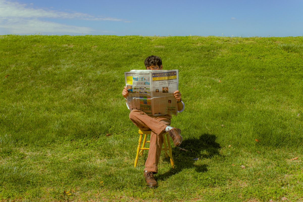A person sitting alone on a chair in a field, reading a newspaper